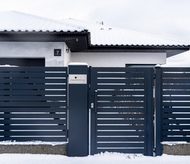 A modern gate with a letterbox and a wireless card reader, mounted in an anthracite panel fence, covered with snow.