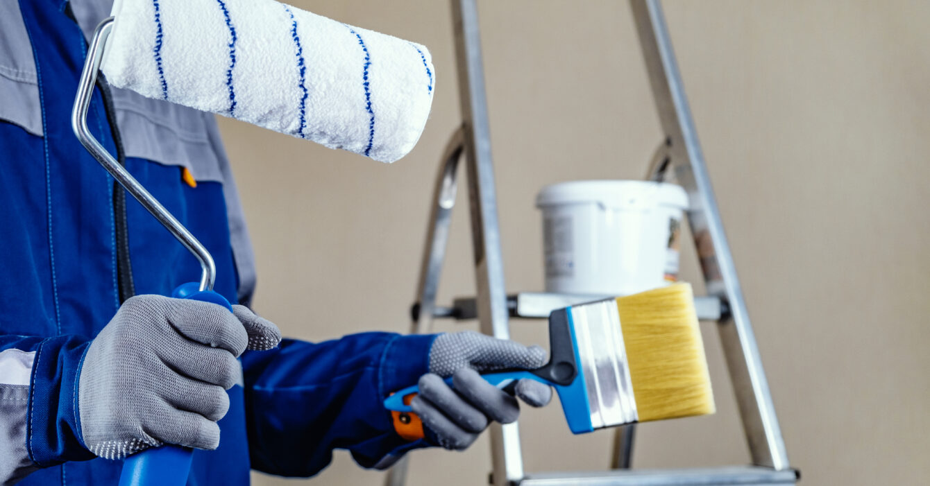 The plaster painter is ready to paint the wall. In the hands of a roller and brush. A stepladder and a bucket of paint in the background. The plaster painter is ready to paint the wall. In the hands of a roller and brush. A stepladder and a bucket of paint in the background.