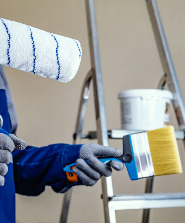 The plaster painter is ready to paint the wall. In the hands of a roller and brush. A stepladder and a bucket of paint in the background.
